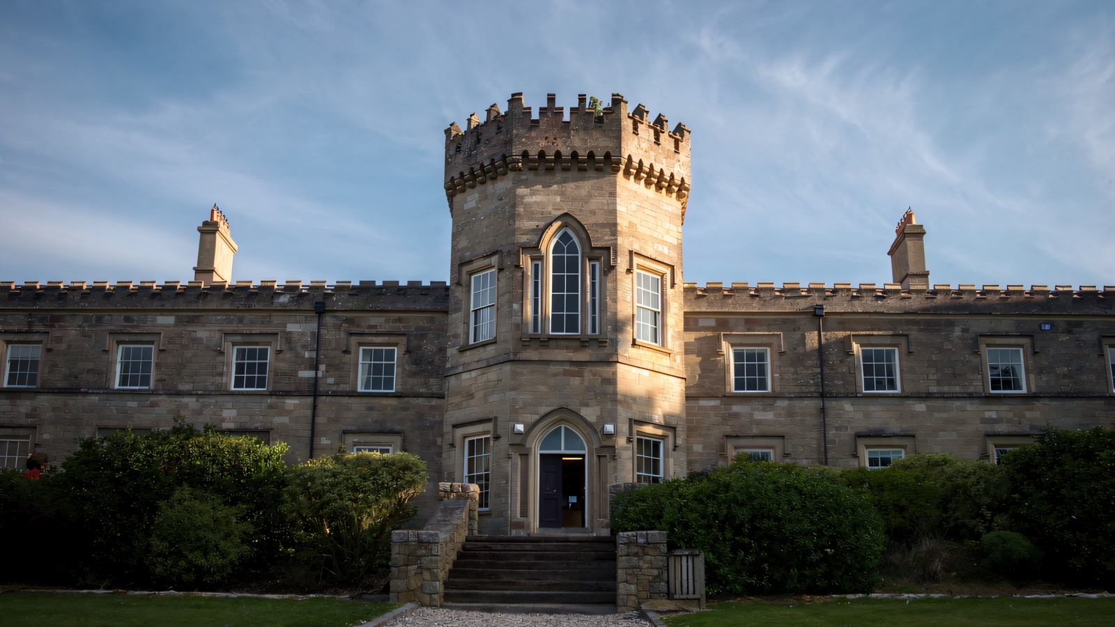The battlemented central tower and grey-stone front of Dungiven Castle above a gravel path and lawn