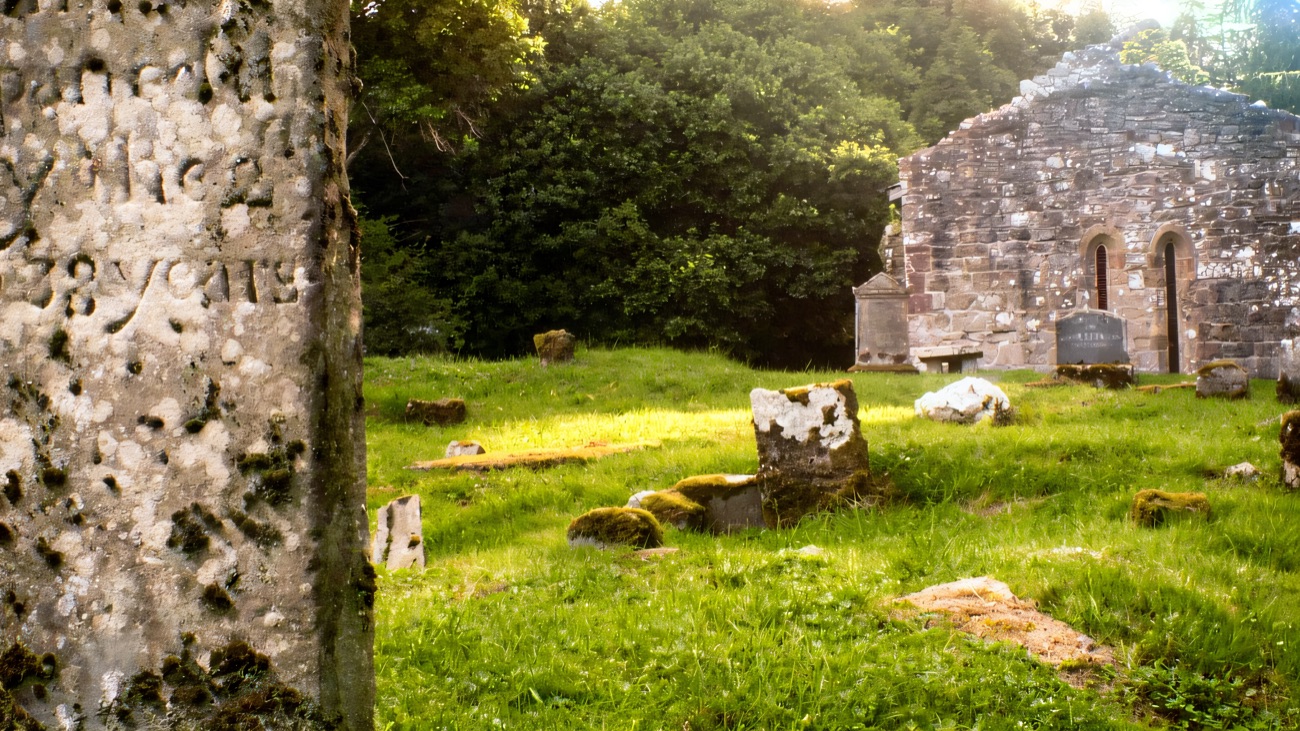 An old inscribed headstone leaning in the long grass before the ruin