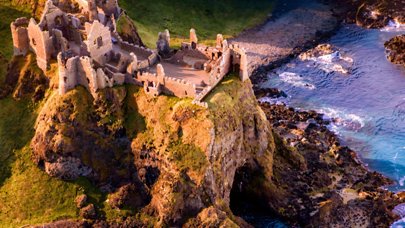Aerial view of Dunluce Castle on its sea-stack with the cove and sea cave below