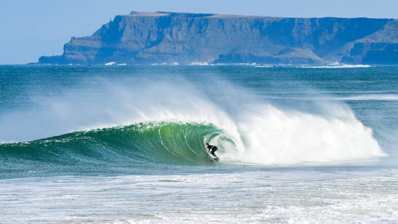 A surfer riding a clean green wave off East Strand with a dramatic headland behind