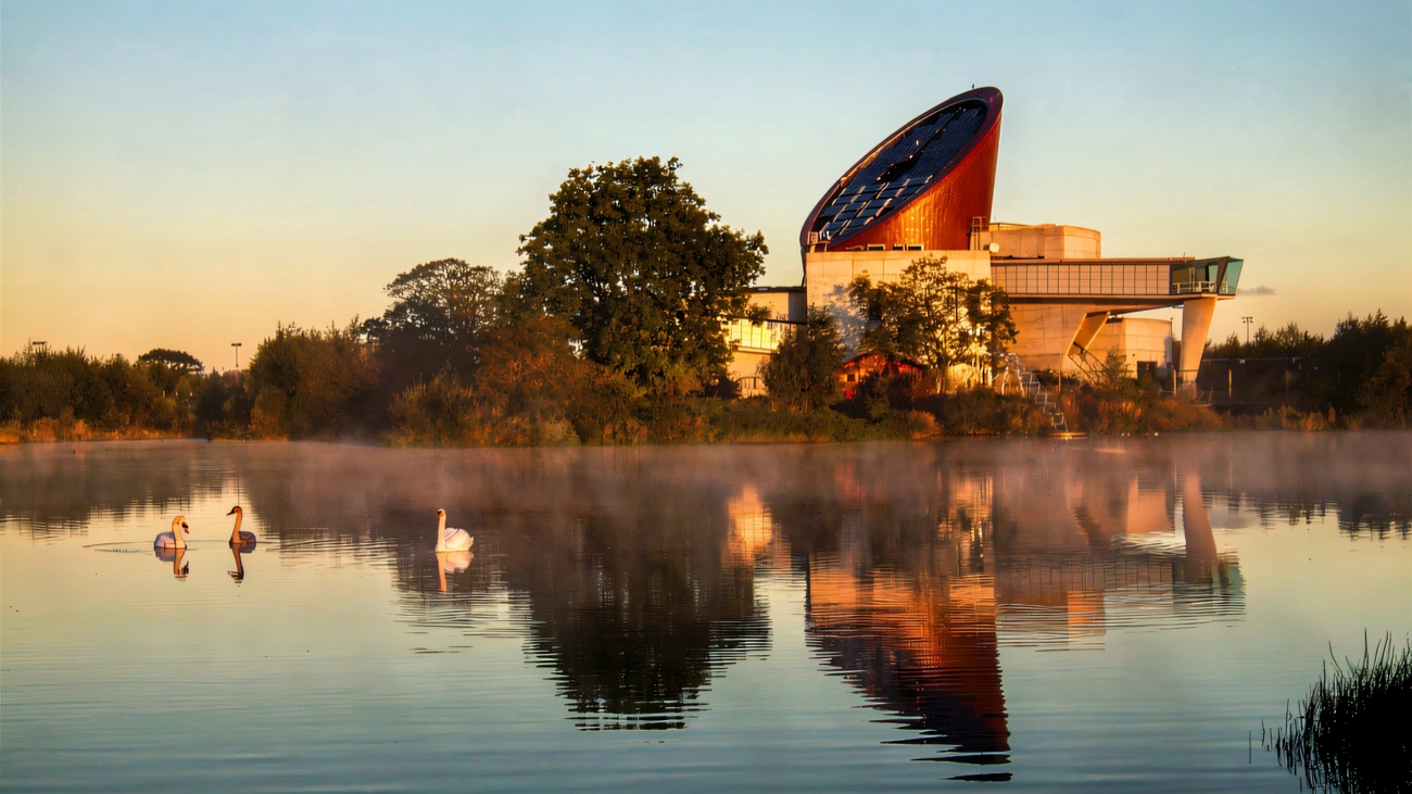 The ECOS Centre and its distinctive solar sail reflected in a misty lake, swans on the water at dawn