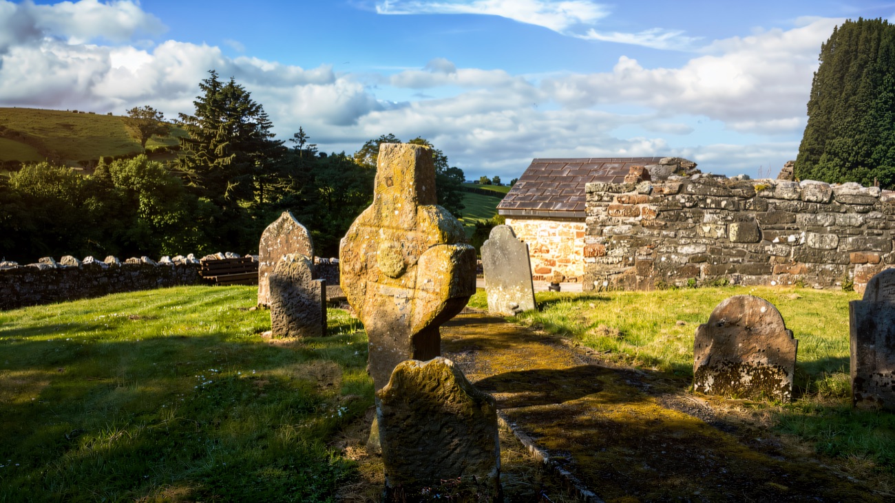 The old ringed stone cross standing among headstones, with the church ruin and green hills behind