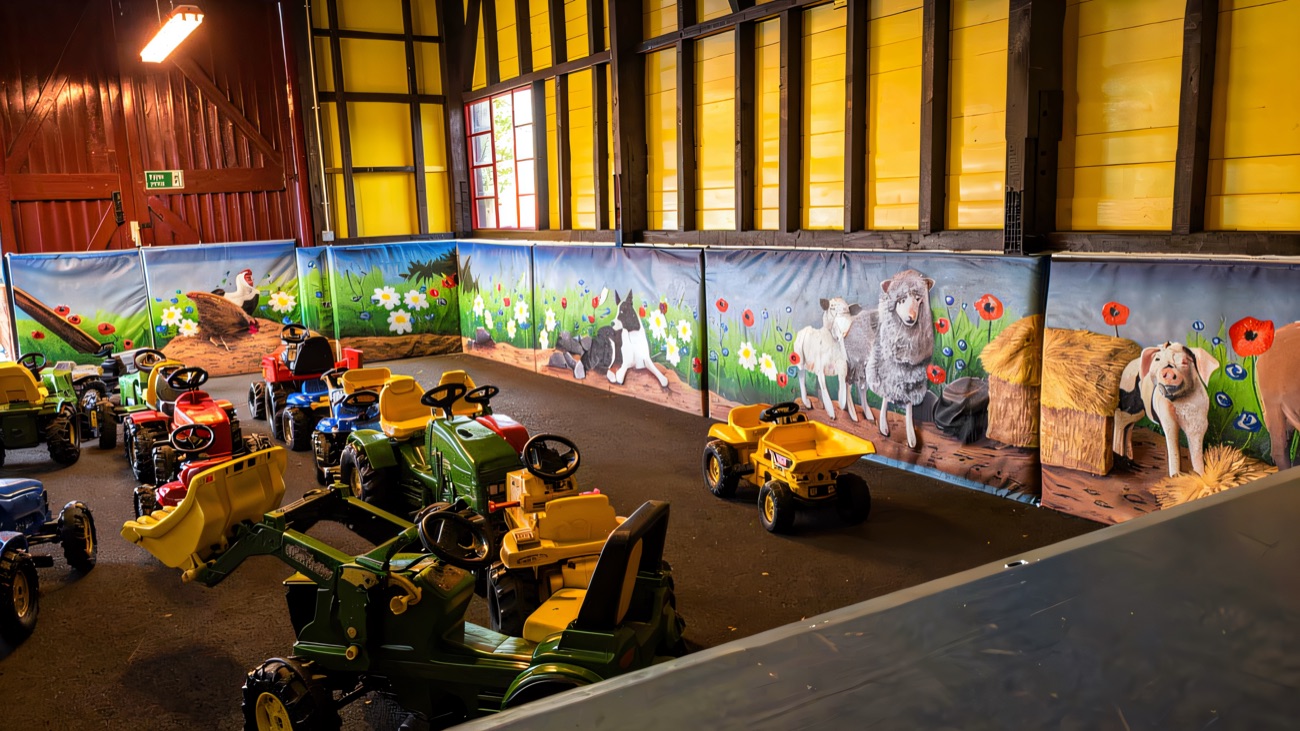 A fleet of ride-on pedal tractors in the indoor play barn with painted farm-animal murals along the walls