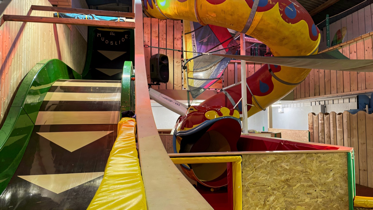 A green wave slide beside a red-and-yellow spiral dragon tube slide inside the indoor play barn