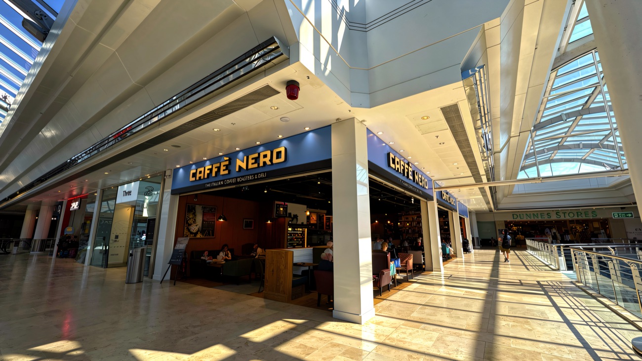 Caffè Nero and the entrance to Dunnes Stores on the bright upper mall at Foyleside, sunshine pouring through the glass roof