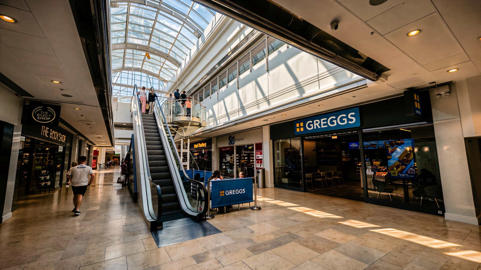 Escalators rising to the upper shopping level under the arched glass roof at Foyleside, with Greggs and The Body Shop either side of the sunlit mall