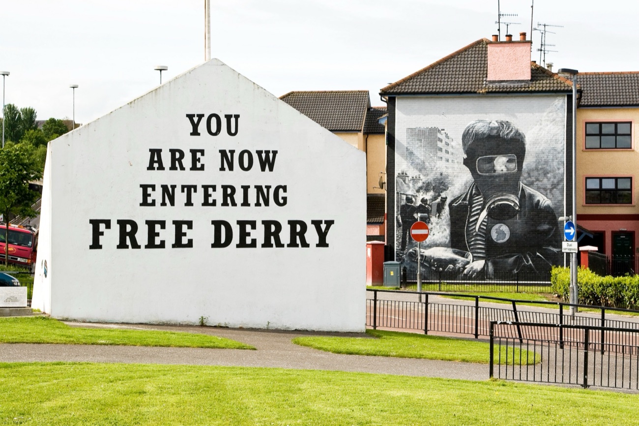 The Free Derry Corner wall in black lettering on white, with a large black-and-white mural on a house gable to the right