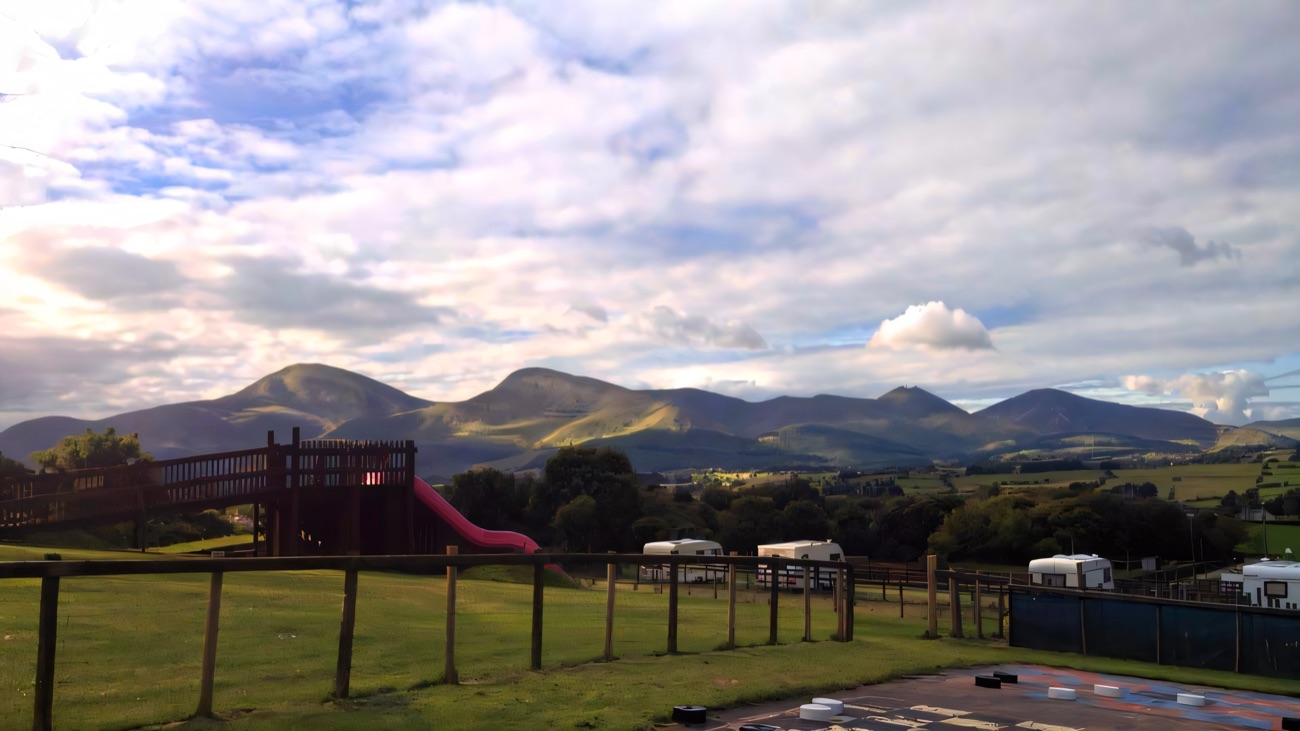 A wooden play tower with a long pink slide in a fenced grass field, with the Mourne Mountains rising behind