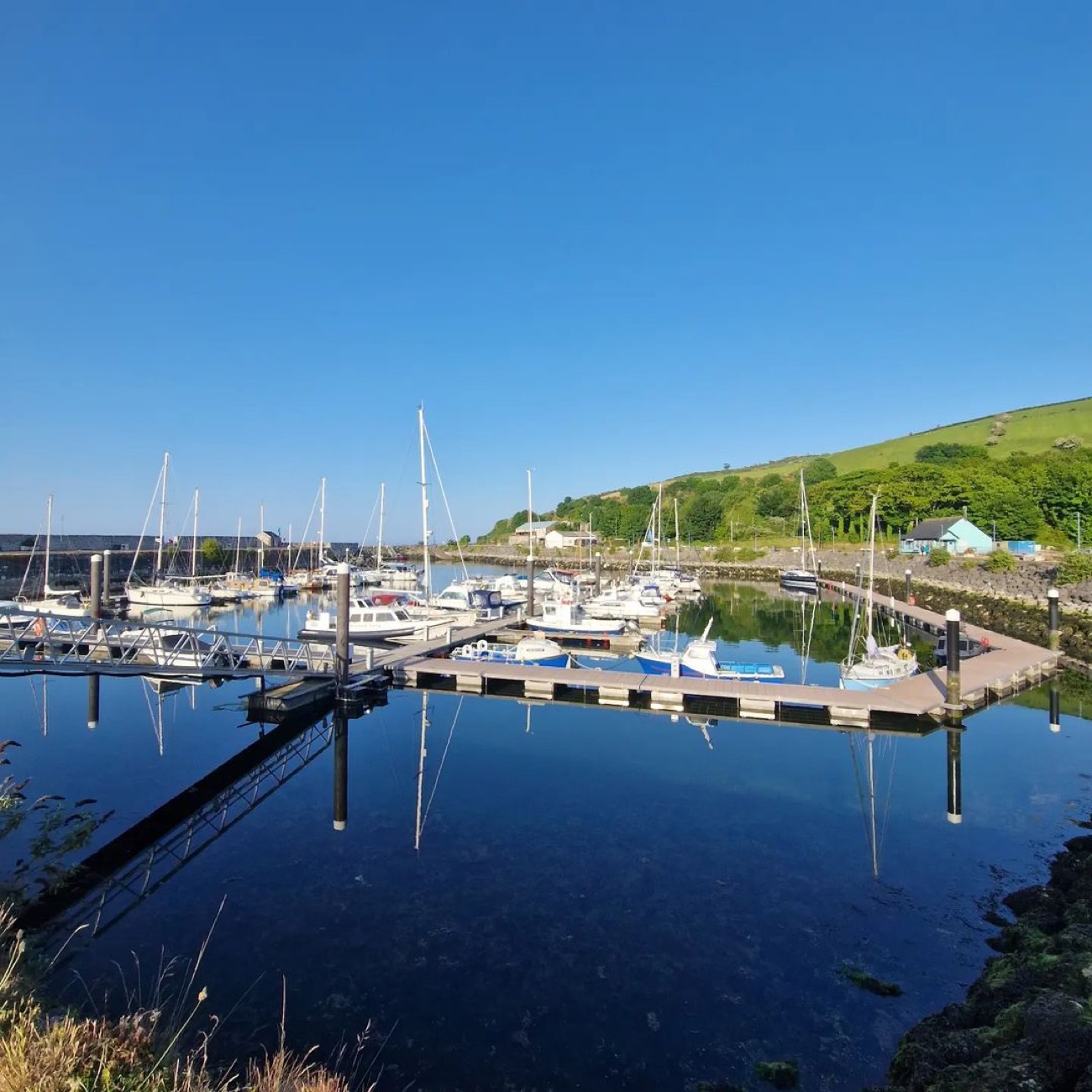 Sailing boats and pontoons in Glenarm marina under a deep blue sky, with a green hill of the Glens of Antrim rising behind