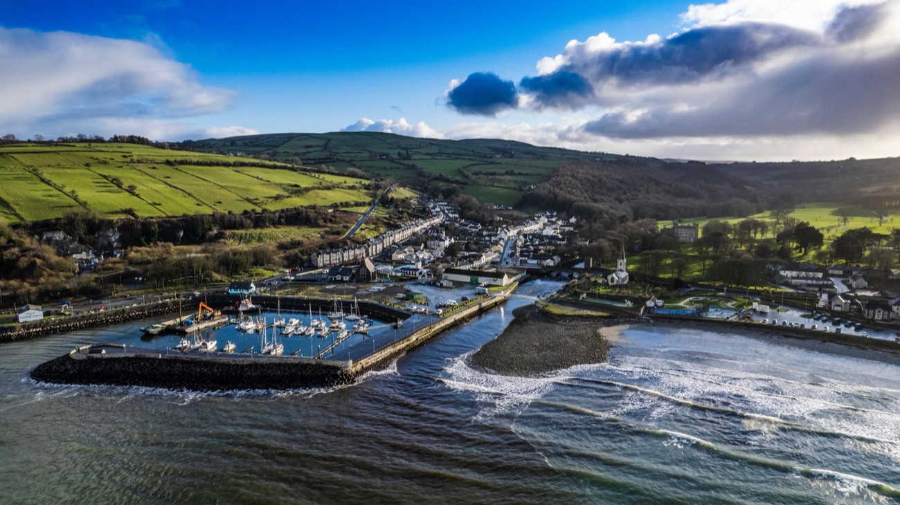 Aerial view of Glenarm harbour and pebble beach curving away with the village and the Glens of Antrim under a dramatic sky