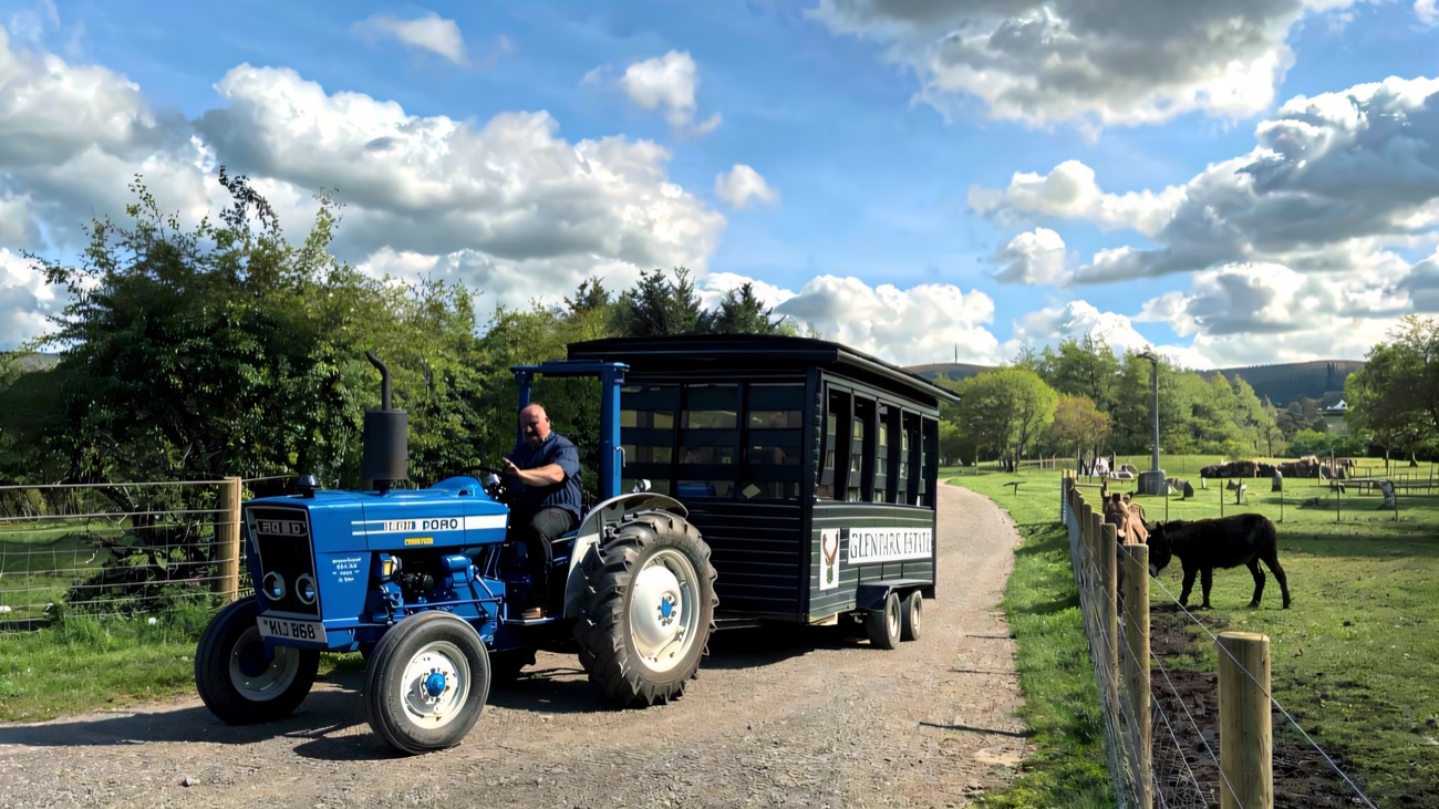 A blue tractor pulling a covered visitor trailer along the farm track, with a pony grazing in the field and the Sperrin hills behind