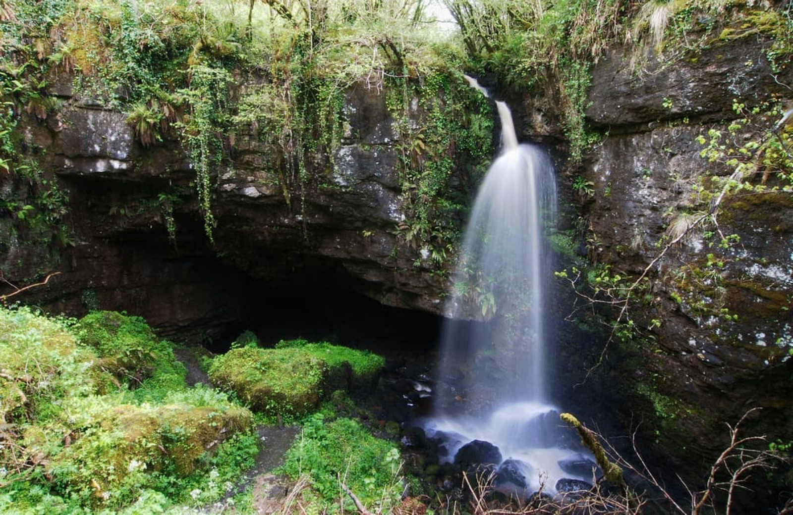 The dark mouth of Pollnagollum Cave under a mossy limestone cliff with a waterfall cascading down beside it