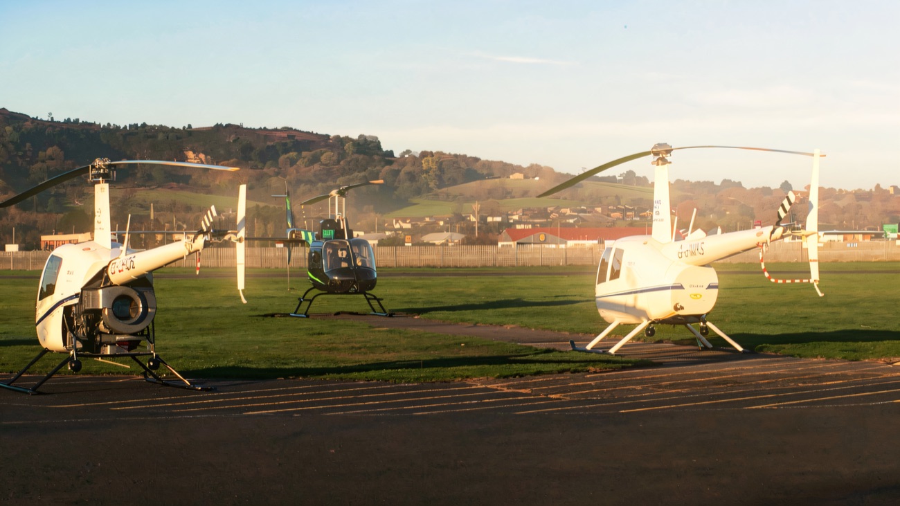Three helicopters parked on the apron on a clear morning, with wooded hills rising behind the airfield