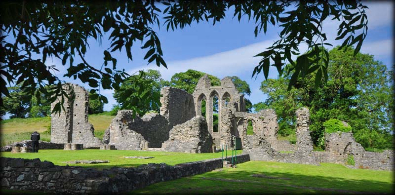 The wide ruins of Inch Abbey across the grass, framed by trees under a blue sky