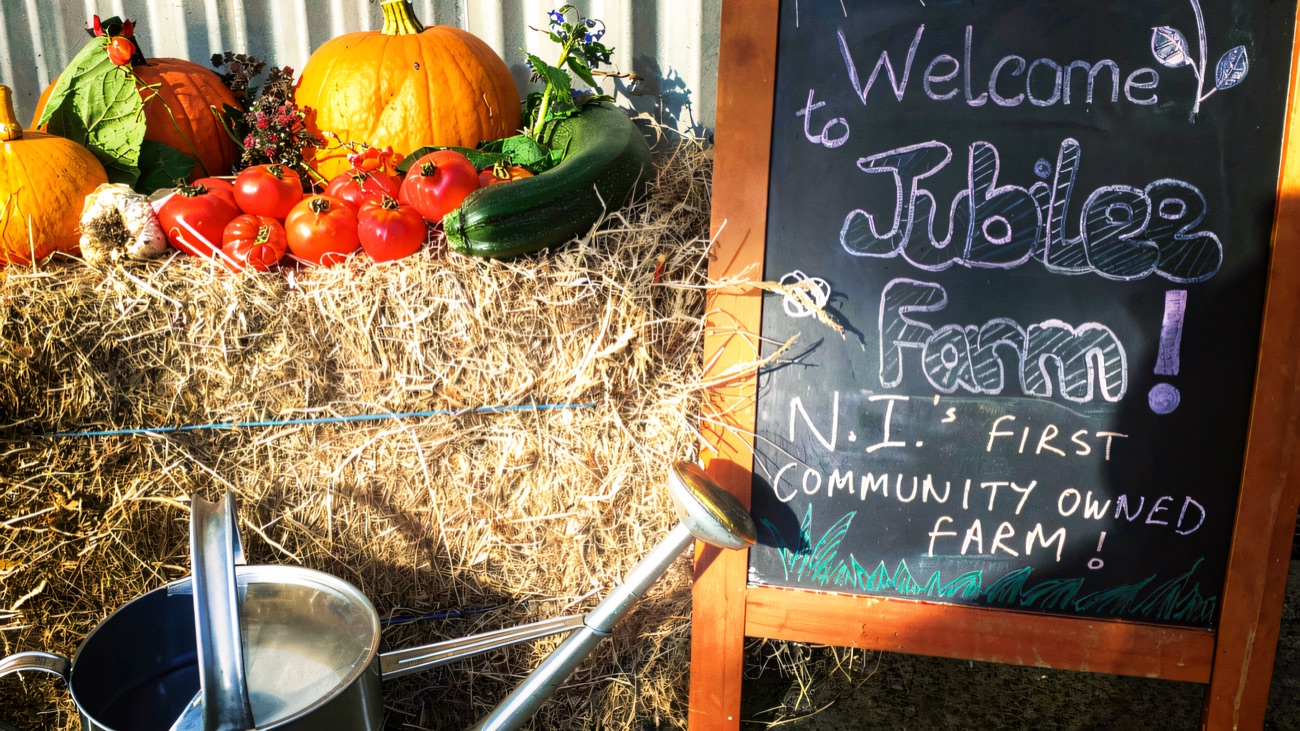 A chalkboard reading Welcome to Jubilee Farm, NI's first community owned farm, beside a hay bale topped with pumpkins and tomatoes