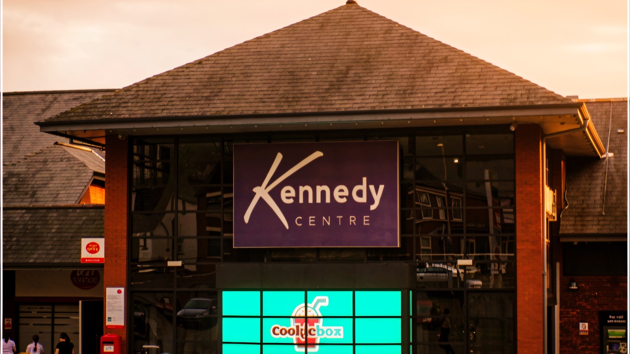 The Falls Road entrance to the Kennedy Centre, with the big purple Kennedy Centre sign above the glass front and red-brick gable