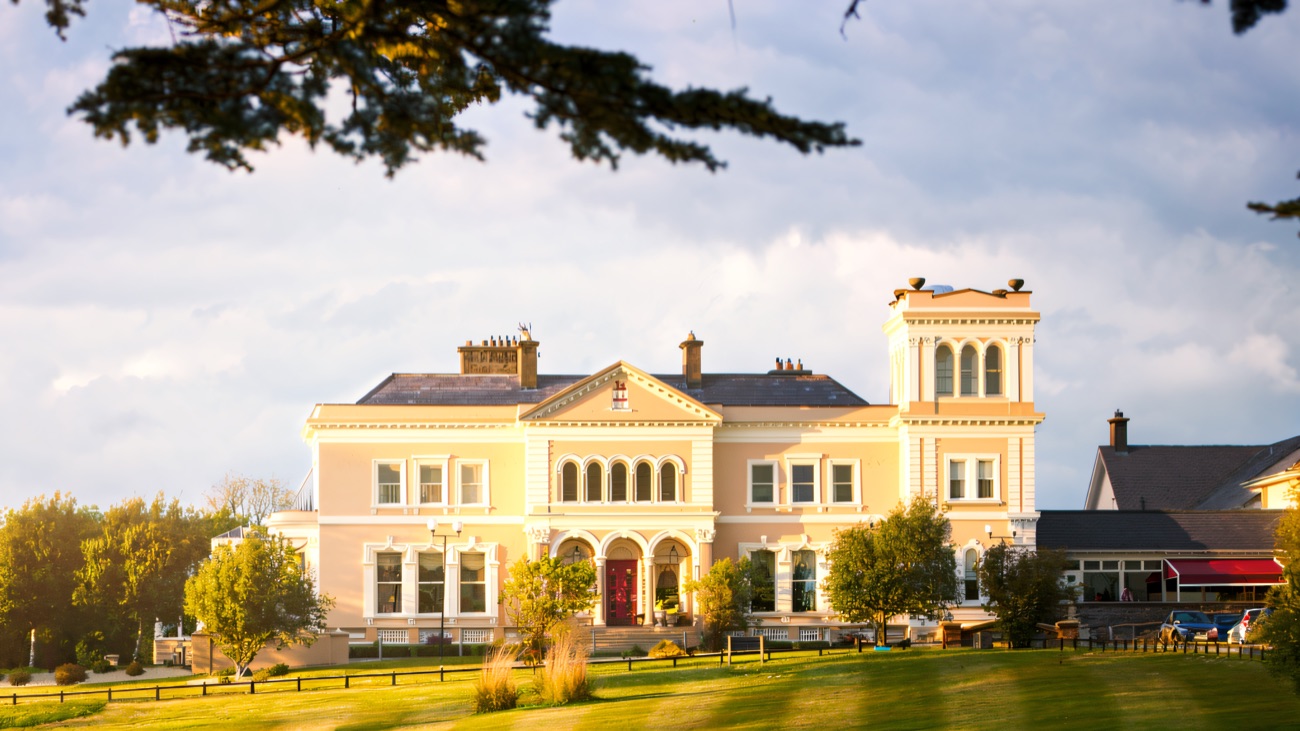 The cream front of the manor hotel seen across a sweeping lawn under a blue sky