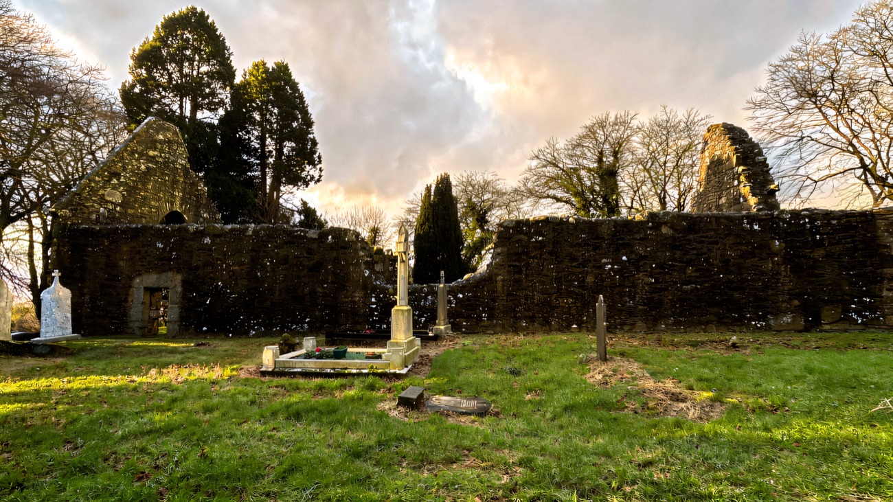 A wide view of the twin ruined churches and their graveyard cross under a spring sky.