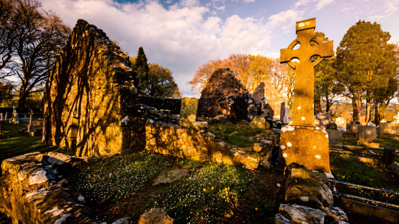 Wildflowers and a Celtic cross catch the sun beside the ancient church walls.