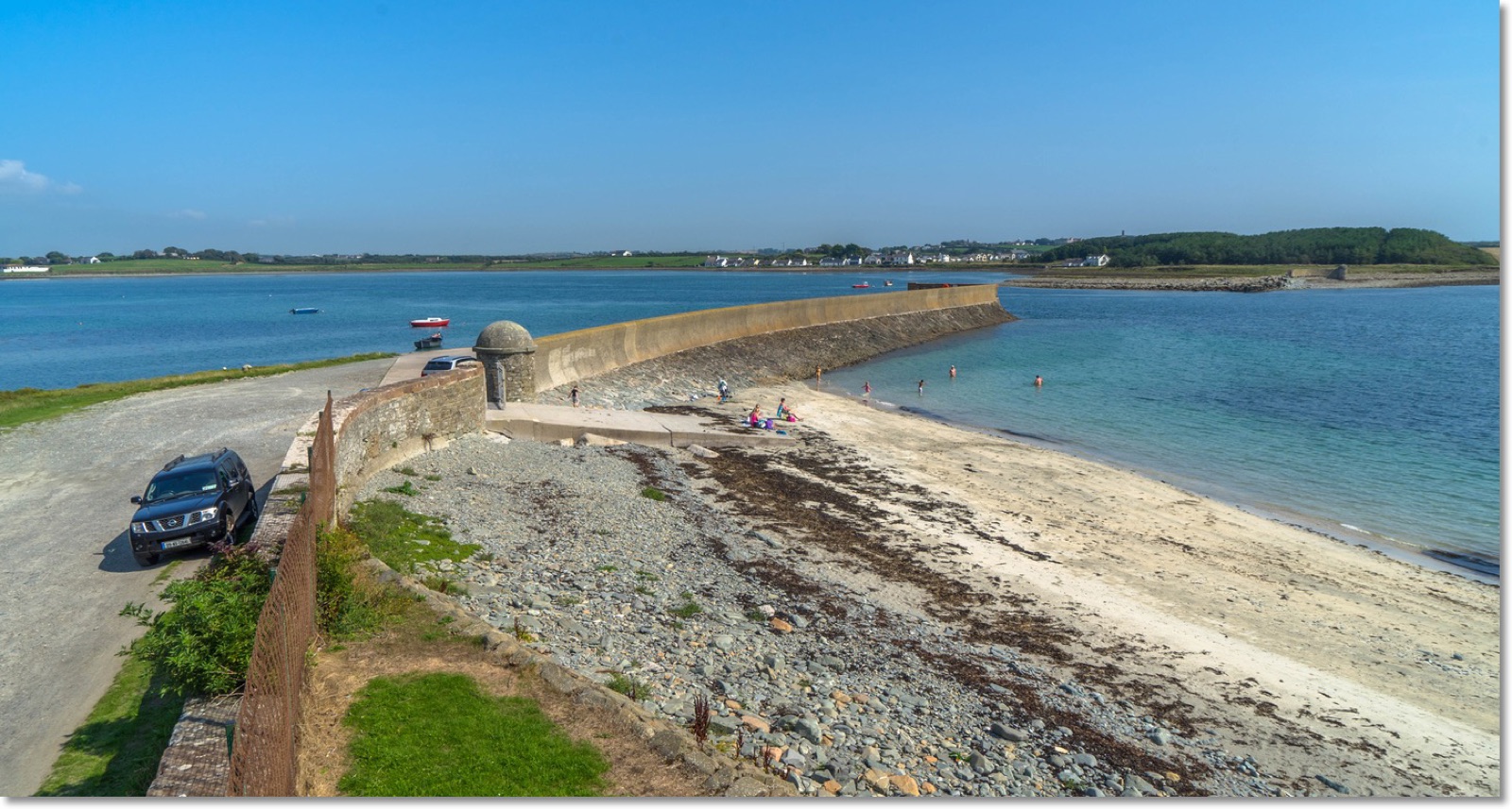 The curving stone harbour pier at Killough reaching out into Coney Island Bay, with a sandy shingle beach in the foreground and the village across the calm blue water under a clear sky