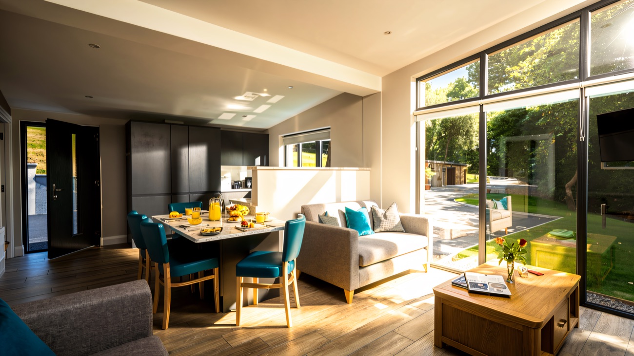 A bright open-plan chalet interior with a kitchen, dining table and sofa opening onto floor-to-ceiling windows and the garden