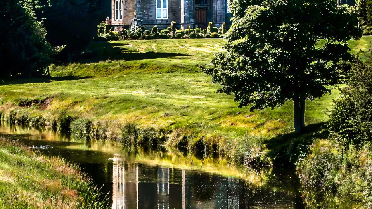 Killymoon Castle reflected in the calm water of the Ballinderry River, framed by a grassy bank and a single tree
