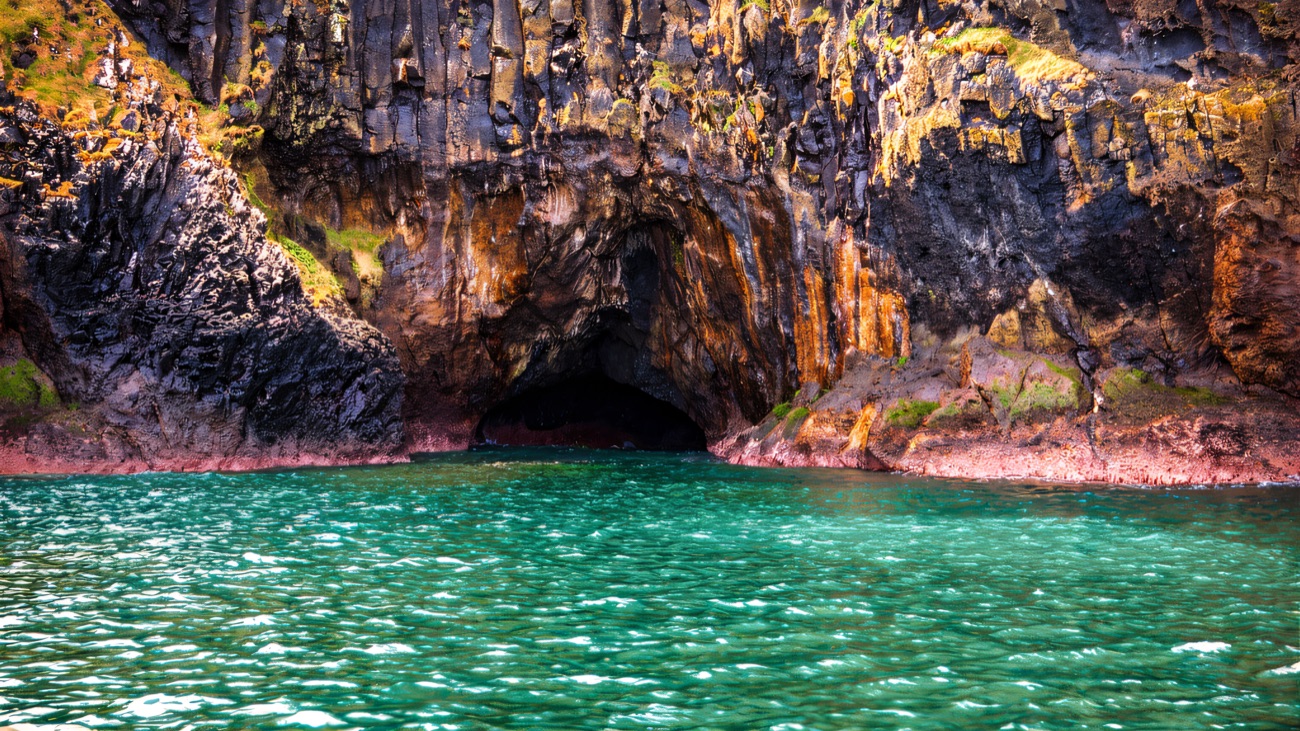 The boat's-eye view of a dark sea cave set into rust-and-black cliffs, jade water at its mouth
