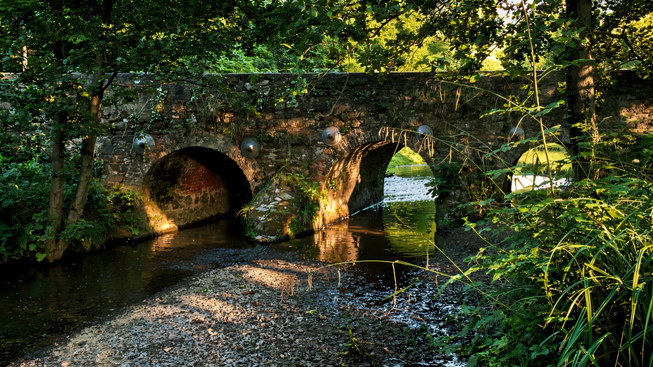 A weathered stone arched bridge crossing the river beside the path