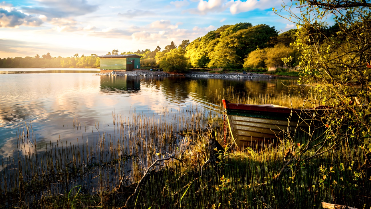 A little wooden rowing boat rests among the reeds, a boathouse across the calm lough