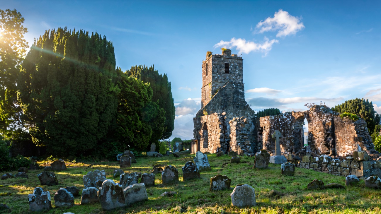 The church ruin and its tower above a graveyard on a sunlit day.