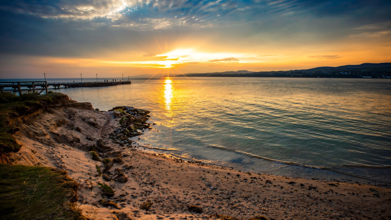 The sun setting over calm water beside a low sandy headland and a long stone jetty