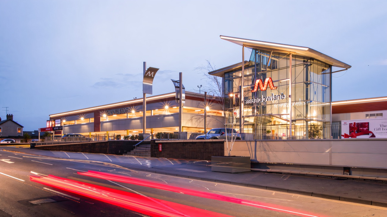 The glass frontage of Meadowlane at dusk with car-light trails along the Moneymore Road and Dunnes Stores lit up inside