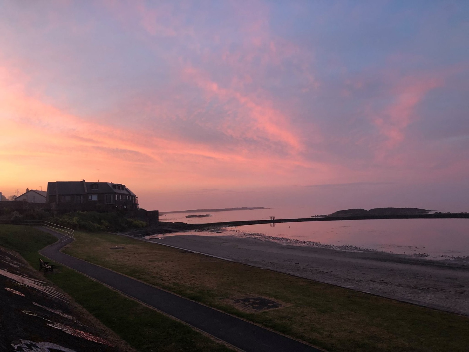 The promenade path, grassy foreshore and sandy beach at Millisle with the seawater lagoon wall curving into a calm sea under a pink dawn sky