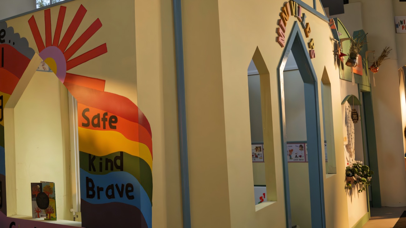 The pale yellow playhouse with its bell tower and a Safe, Kind, Brave rainbow mural