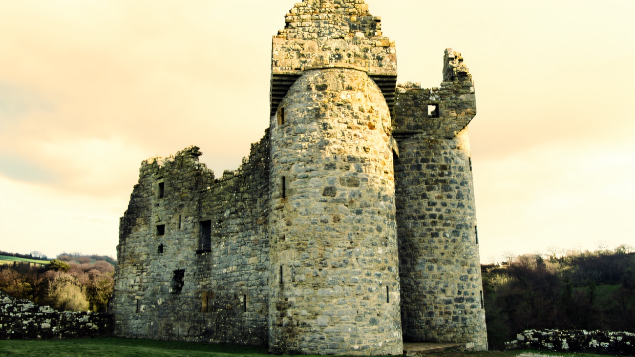 The two tall round entrance towers of Monea Castle head-on, with the ruined shell and green setting