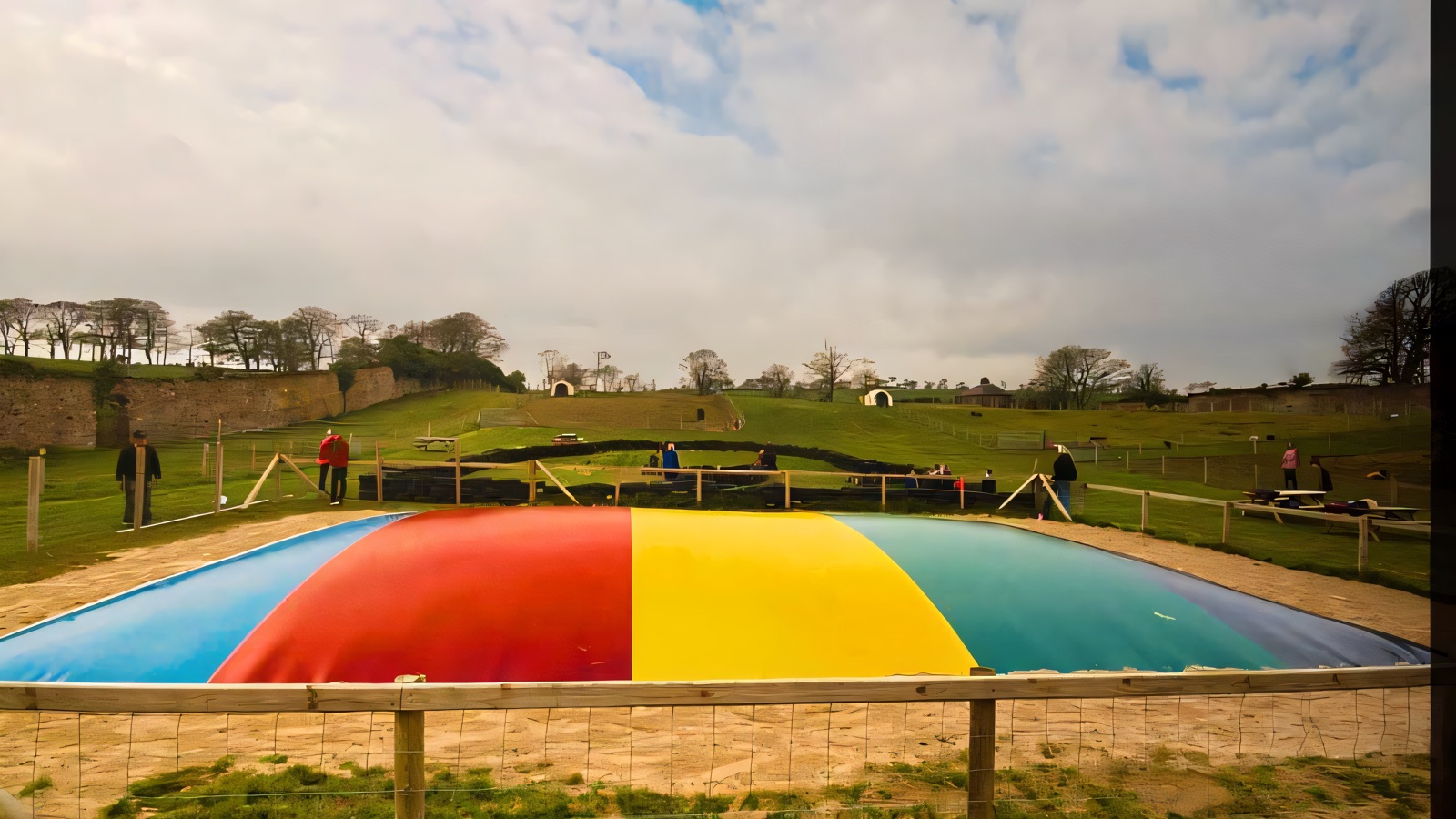 The big rainbow jumping pillow set in sand at Mountpanther Farm Park, with rolling green hills and the deer field behind