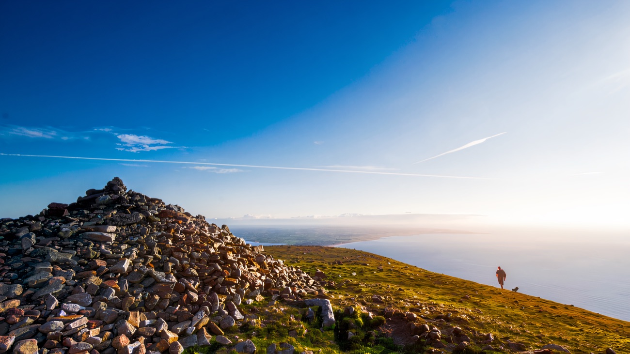 A lone walker at the summit cairn with the sea spread out far below