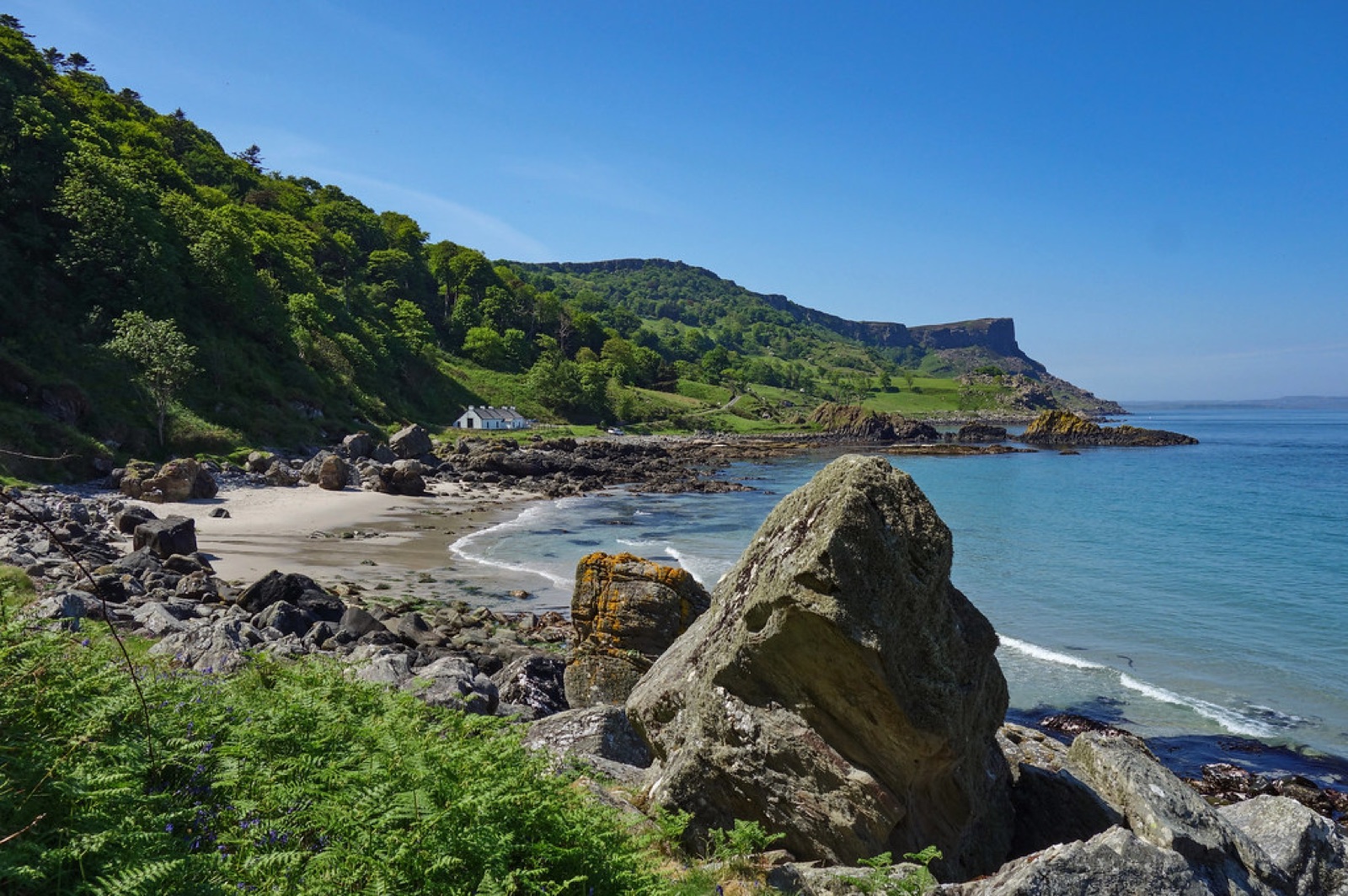 Murlough Bay from the shore — a white cottage below green slopes with the Fair Head cliff rising to the right above turquoise sea