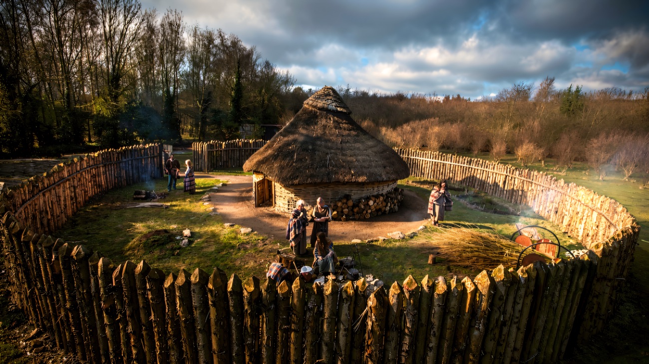 The reconstructed Iron Age roundhouse inside its timber palisade, with interpreters at work.