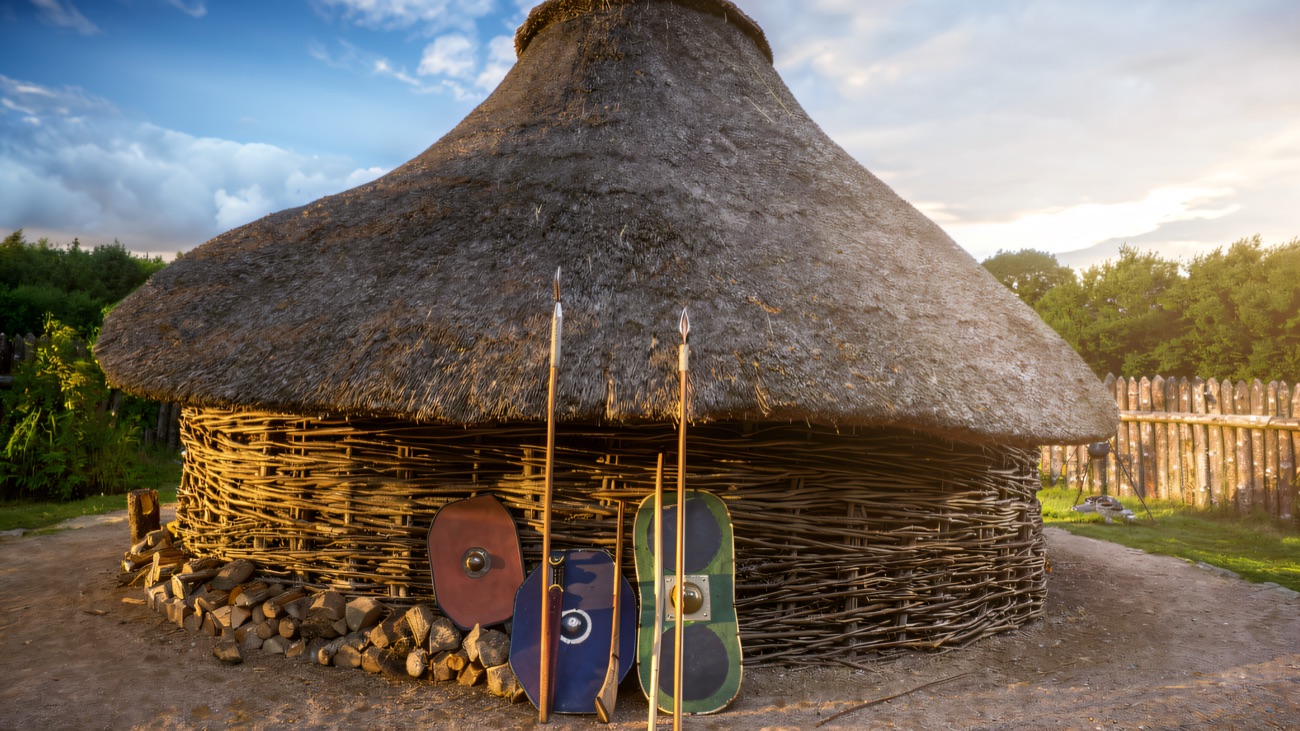 The thatched roundhouse close up, with painted shields and spears leaning at the door.