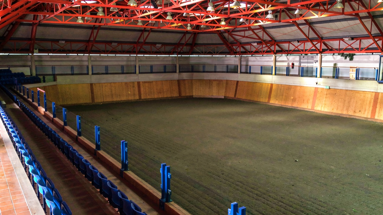 The large indoor arena at Necarne equestrian centre, red steel roof and tiered blue spectator seating around a sand surface