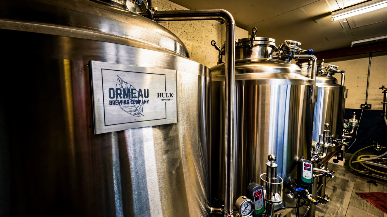 Stainless steel brewing tanks lined up inside the brewhouse, with the Ormeau Brewing Company plate fixed to the front vessel.