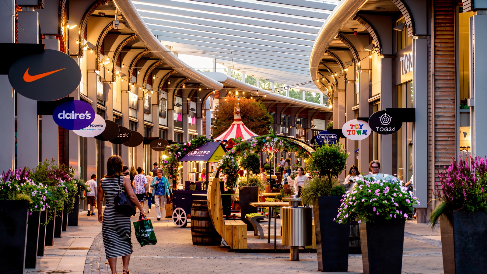 The open-air mall street at The Boulevard Banbridge, with curved canopy walkways over the shopfronts, planters down the middle and shoppers strolling between the stores