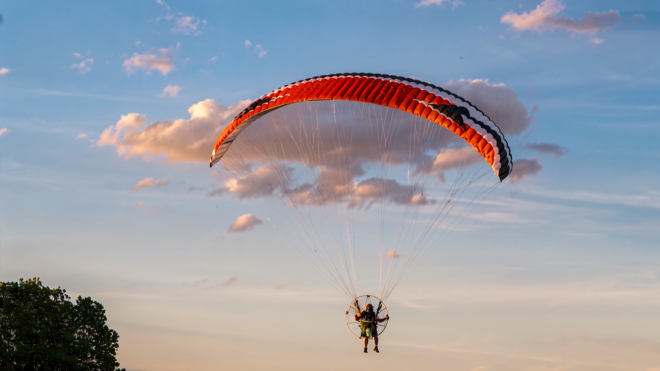A red and white paramotor wing climbing into a blue sky above the treeline
