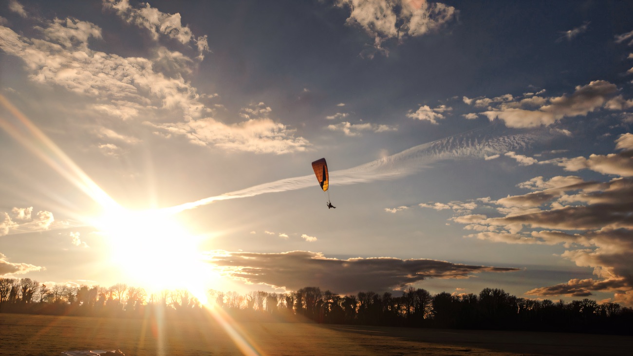 A paramotor flying high over open farmland as the low sun breaks through