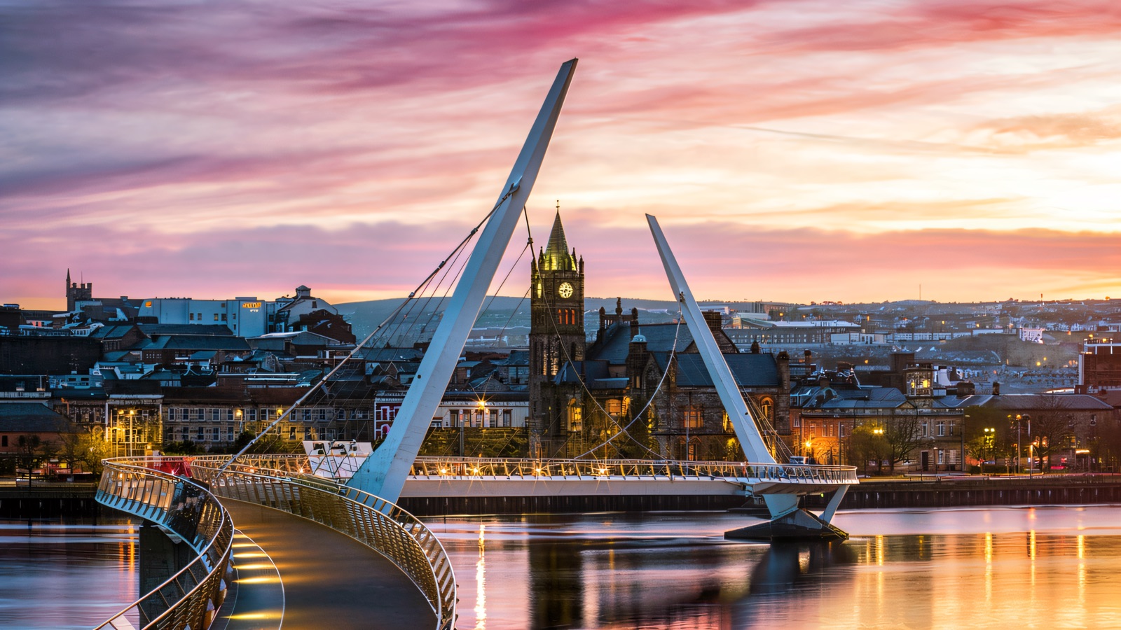 The white cable-stayed Peace Bridge curving across the River Foyle at pink dusk with the Guildhall and Derry city lights behind