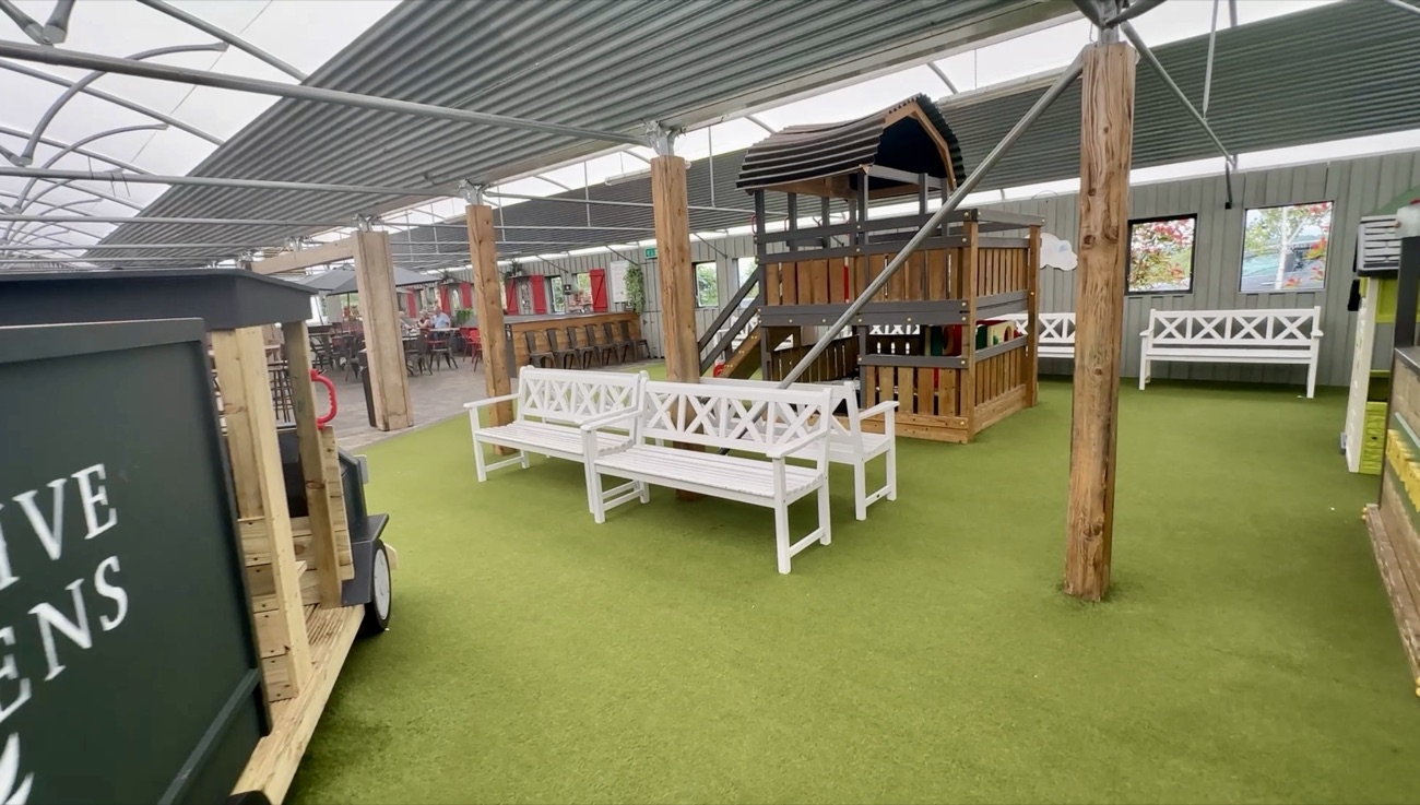 A wide view of the play area with a wooden playhouse and benches, the garden-centre café seating visible in the background
