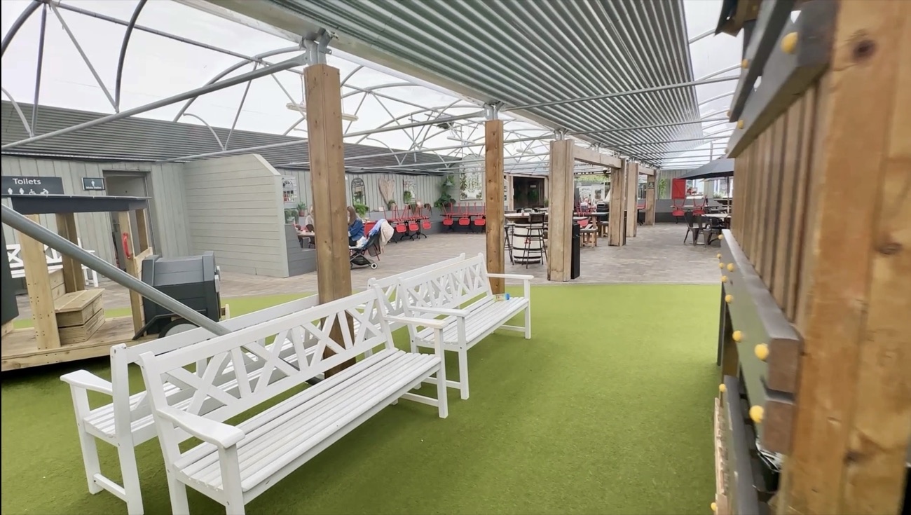 White benches on the astroturf beside the play structures, looking through to the café tables and toilets