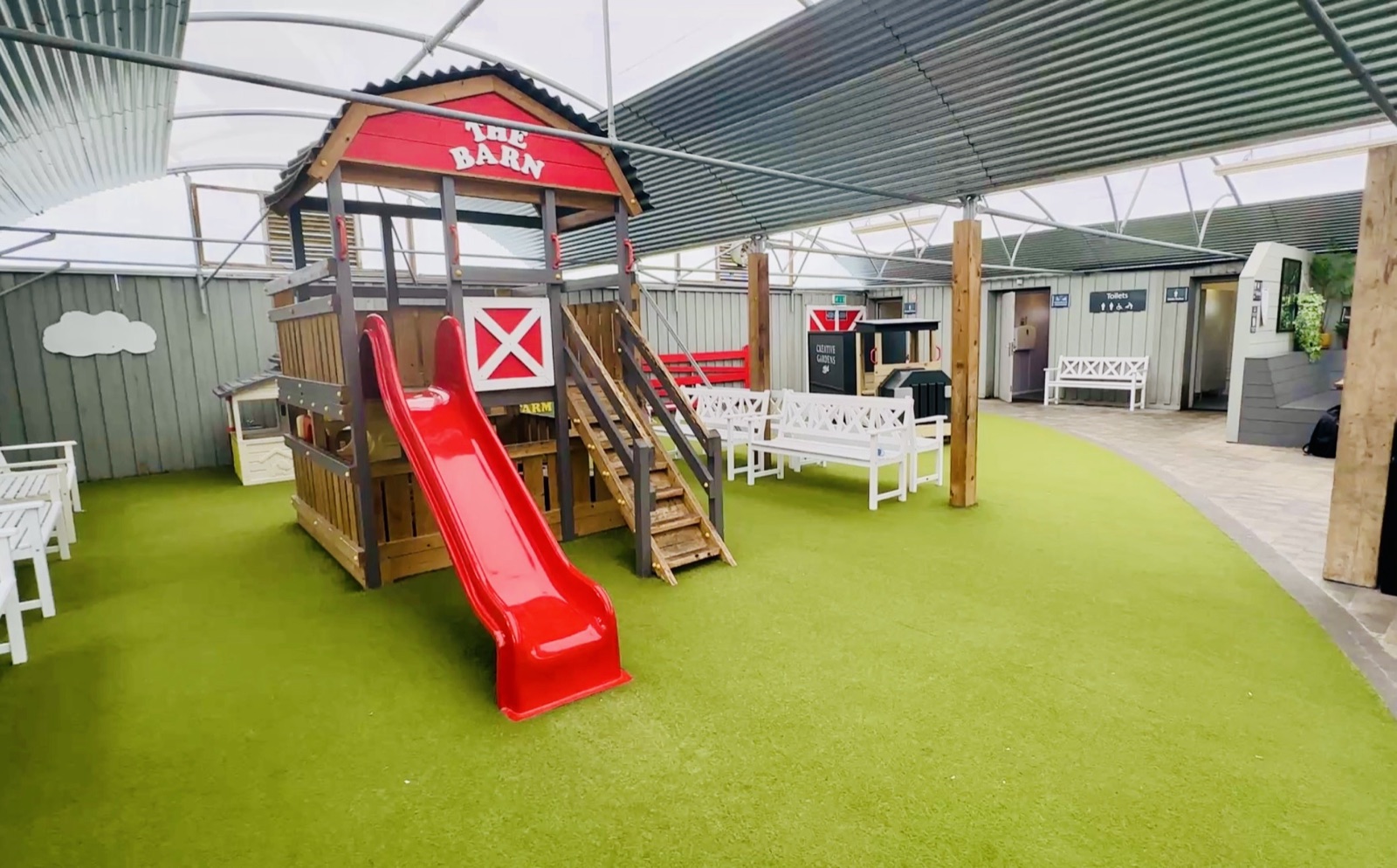 The covered play zone at Creative Gardens Bushmills, with 'The Barn' wooden playhouse, a red slide and white benches on bright green astroturf under a polytunnel canopy