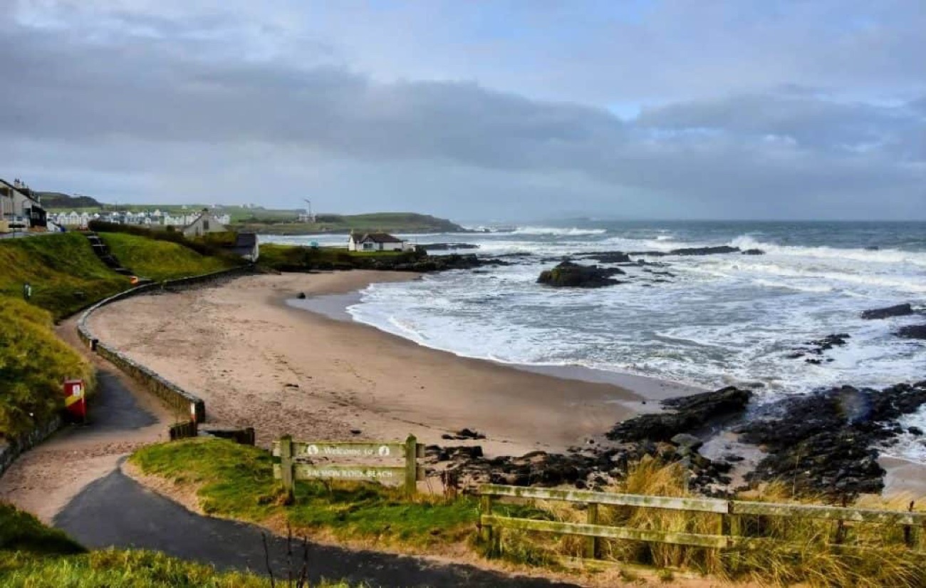 The sandy Salmon Rock beach at Portballintrae with a welcome sign, rocks and waves rolling in
